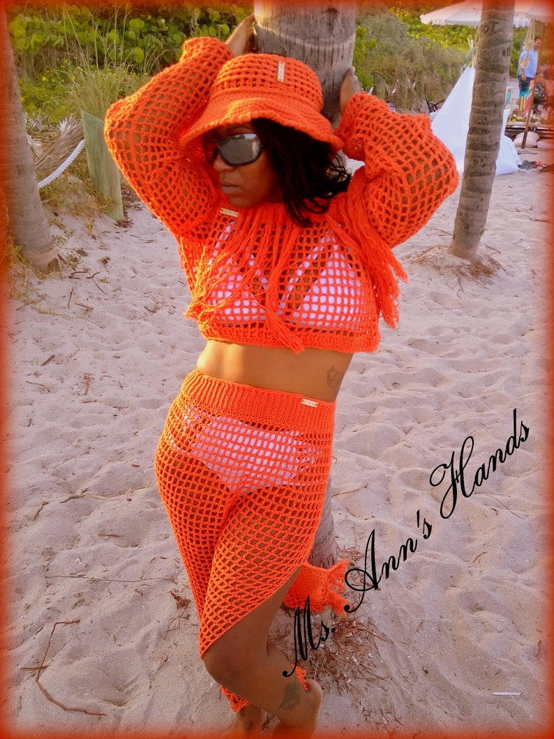 May include: Orange crochet top and pants set with fringe detailing. The top is a cropped style and the pants are wide-leg. The model is wearing a white crocheted hat and sunglasses. The text "Mrs. Ann's Hands" is visible in the image.