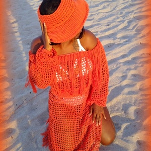 May include: A woman wearing a bright orange crochet dress and matching sun hat. The dress has a loose fit and features a fringe trim. The hat is wide-brimmed and has a crocheted design.
