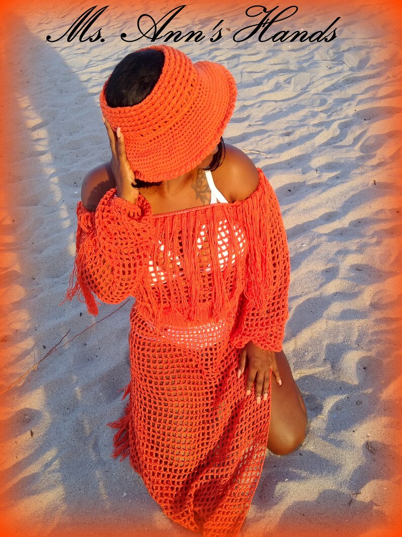 May include: A woman wearing a bright orange crochet dress and matching sun hat. The dress has a loose fit and features a fringe trim. The hat is wide-brimmed and has a crocheted design.