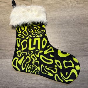 Könnte beinhalten: Schwarzer Weihnachtsstrumpf mit neongrünem, abstraktem Muster und dem Wort "VOLTO". Die Oberseite des Strumpfes hat eine weiße Kunstfellborte und eine schwarze Schlaufe zum Aufhängen. Eine festliche Weihnachtsdekoration.