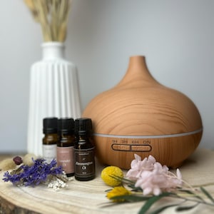 Könnte beinhalten: Ein Holz-Diffusor für ätherische Öle mit einer hellen Holzoptik und einem weißen Bedienfeld mit dem Text "1H 3H 6H ON LIGHT MIST". Im Vordergrund befinden sich drei kleine Glasfläschchen mit ätherischem Öl, die mit "Lavender Oil" und "Citronengras Oil" beschriftet sind.