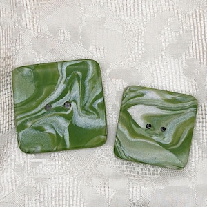 May include: Two square green buttons with a marbled white pattern. Each button has two holes for sewing. The buttons are a light green color with a swirled white design. The buttons are made of a glossy material.