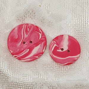 May include: Two round buttons with a marbled design in shades of pink and white. Each button has two small holes for sewing. The buttons are different sizes, with a larger one and a smaller one.