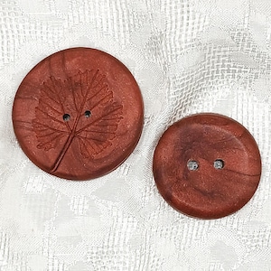 May include: Two round, copper-colored buttons with a metallic finish. The larger button features an embossed leaf pattern and two central holes. The smaller button is plain with a smooth surface and two central holes for attachment to fabric.
