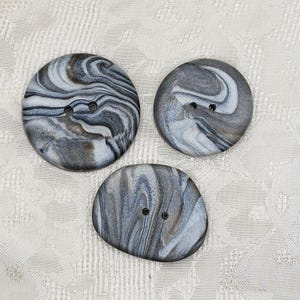 May include: Three round buttons featuring a marbled pattern of swirling gray, black, and white tones. Each button has two small holes in the center for attachment. The buttons vary slightly in size and shape, displaying a smooth, polished surface.