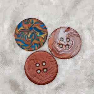 May include: Three round buttons with four holes each. One button features a marbled pattern of blue, gold, and brown. Another button has a white and copper marbled swirl design. The third button is solid brown with a textured leaf-like pattern.