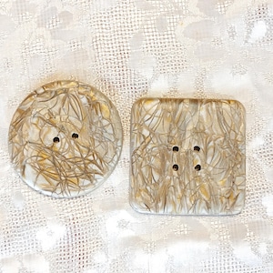 May include: Two decorative buttons, one round and one square, with a cream and gold abstract design. The round button has two holes, and the square button has four holes. The buttons are set against a textured white background.
