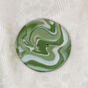 May include: A large, round button with a marbled design in shades of green and white. The button has two small holes for sewing and is set against a textured, off-white background. This button is suitable for clothing and craft projects.