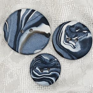 May include: Three circular buttons of varying sizes featuring a marbled pattern in shades of black, white, and grey. Each button has two holes in the center for attachment. The surface of the buttons displays a smooth, swirling, and fluid design.
