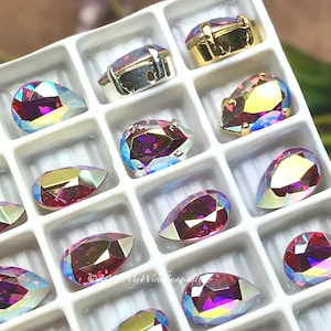 Puede incluir: Una colecci&oacute;n de pedrer&iacute;a iridiscente en forma de l&aacute;grima en una bandeja blanca dividida. Las piedras brillan con colores rosa, morado y dorado. Algunas est&aacute;n engastadas en engastes de metal plateado o dorado.
