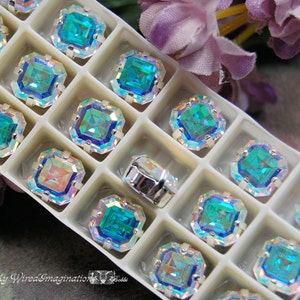 May include: A white plastic tray with 12 compartments filled with iridescent blue square-shaped rhinestones. The rhinestones have a rainbow effect.