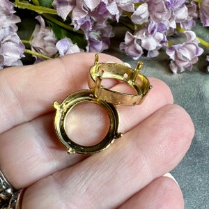 May include: Two gold-toned metal settings for round cabochon stones. The settings have four prongs and a smooth, rounded edge.