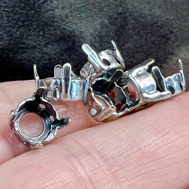 K&ouml;nnte beinhalten: Eine Ansammlung kleiner, silberfarbener Metallfassungen f&uuml;r Schmuck mit kreisf&ouml;rmigen &Ouml;ffnungen und mehreren vertikalen Krappen, die dazu dienen, Steine zu halten. Diese Metallkomponenten haben eine texturierte, oxidierte Oberfl&auml;che.