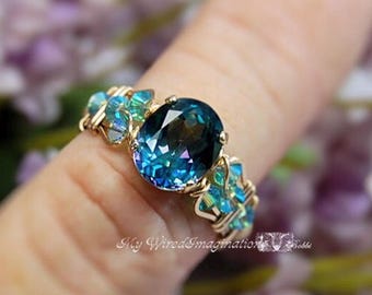 May include: A wire-wrapped ring featuring a large oval-cut dark blue center stone flanked by smaller faceted teal crystals. The band is constructed from thin gold-toned wire, and the text My WiredImagination Bobbi is visible at the base.
