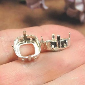 Könnte beinhalten: Zwei silberfarbene Schmuckfassungen aus Metall mit quadratischer Basis und vier aufrechten Krappen zur Aufnahme eines Edelsteins. Die Fassungen haben eine glatte, reflektierende Oberfläche und kleine kreisförmige Aussparungen an den Seitenwänden.