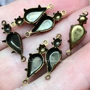 Könnte beinhalten: Sechs antike goldfarbene Metall-Tropfenfassungen mit Zinken und Sprungringen. Die Fassungen sind für das Einsetzen eines Steins oder Cabochons konzipiert.