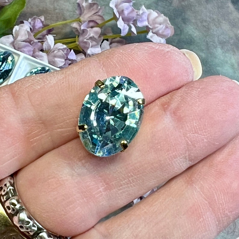 May include: A light blue oval-shaped rhinestone with gold settings. The rhinestone is set on a finger.