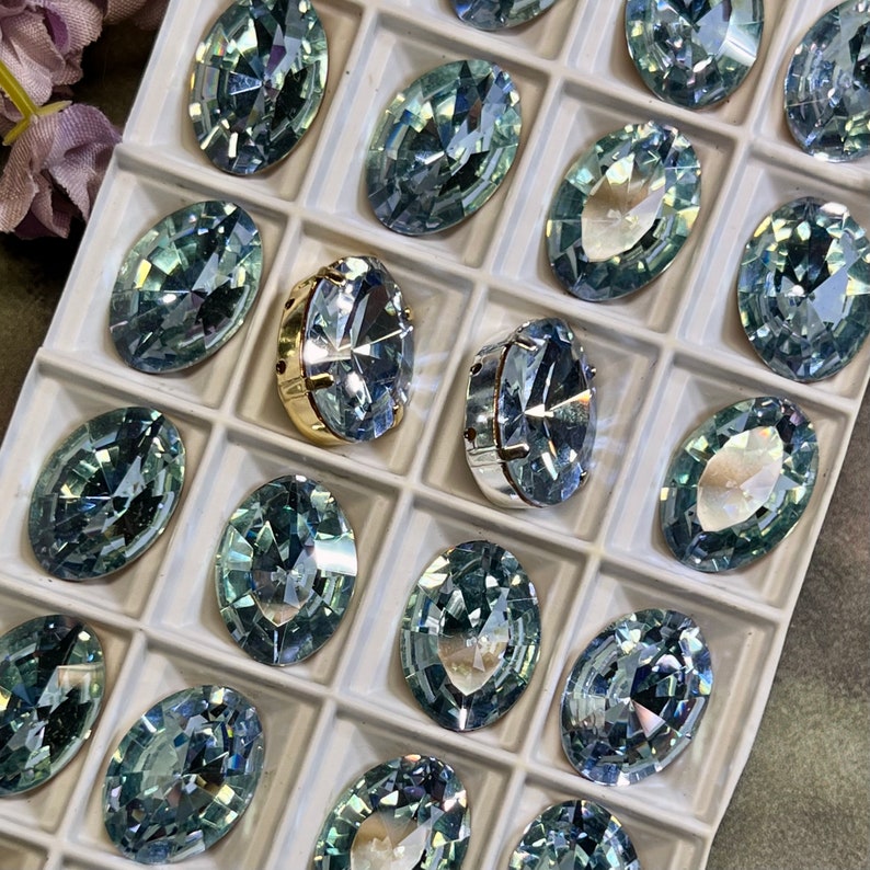 May include: A close-up view of a tray filled with oval-shaped, light blue rhinestones. The rhinestones are arranged in rows and columns, and they have a faceted design that reflects light.