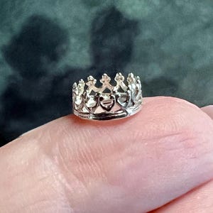 May include: A small, silver-colored metal crown charm featuring a series of pointed arches and heart-shaped cutouts along the band. The crown has a polished, reflective surface and a circular base, designed with intricate detailing throughout the structure.