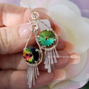 May include: Silver dangle earrings with iridescent crystal pendants and fringe detailing. The crystals reflect a spectrum of colors, including green, yellow, and purple. The earrings are held by a person in front of a soft, blurred background.