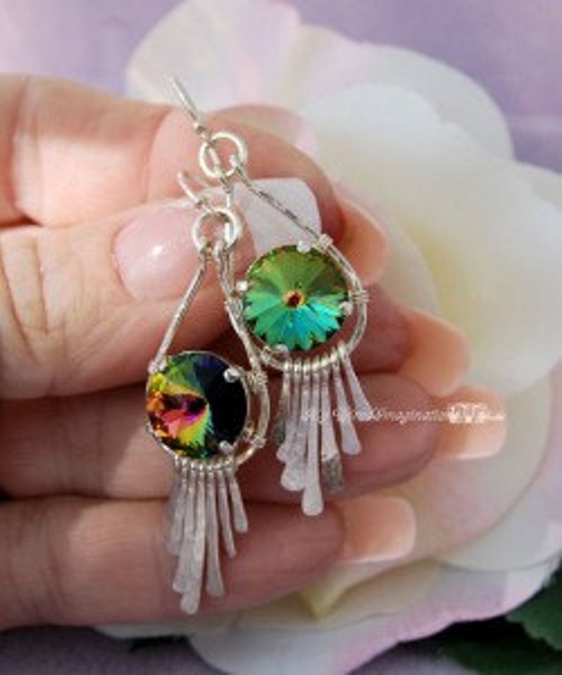 May include: Silver dangle earrings with iridescent crystal pendants and fringe detailing. The crystals reflect a spectrum of colors, including green, yellow, and purple. The earrings are held by a person in front of a soft, blurred background.