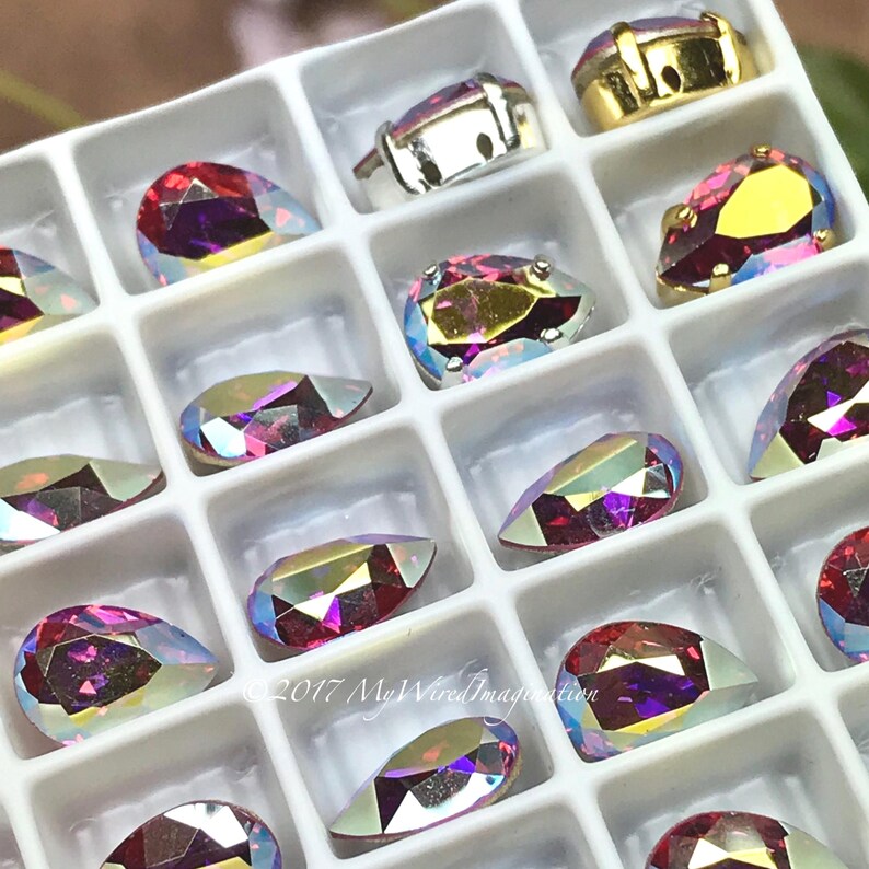 Puede incluir: Una bandeja blanca llena de pedrer&iacute;a iridiscente de varias formas. La pedrer&iacute;a muestra una gama de colores, incluyendo rosa, morado y dorado. Algunos est&aacute;n engastados en engastes de metal plateado o dorado. Se incluyen formas de l&aacute;grima y ovaladas.