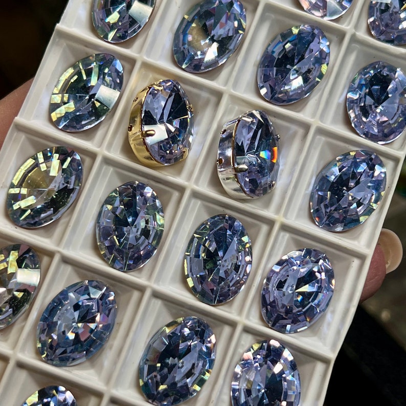 May include: A close-up of a white tray filled with various sizes of oval-shaped, clear, iridescent glass rhinestones. The rhinestones are arranged in rows and columns, with some of the rhinestones having a gold setting.
