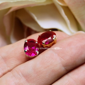 May include: Two round, bright pink, faceted gemstones set in gold settings. The gemstones are on a finger.