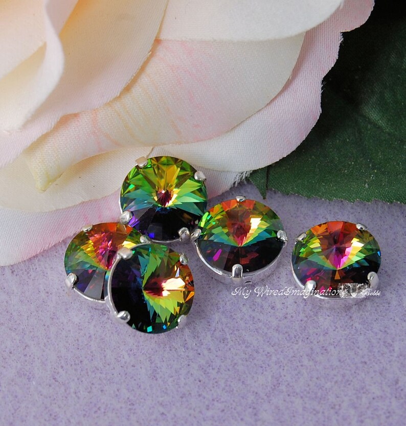 May include: Five iridescent round crystals with silver settings. The crystals display a vibrant spectrum of colors, including green, yellow, orange, and purple. The crystals are arranged on a light purple surface, with a soft pink and white flower in the background.