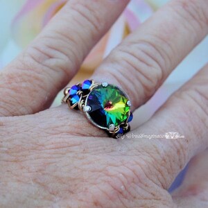 May include: A handcrafted ring featuring a large, iridescent crystal with a rainbow effect. The crystal is set in a silver-toned metal with gold wire accents and smaller blue crystals. The ring is on a finger.