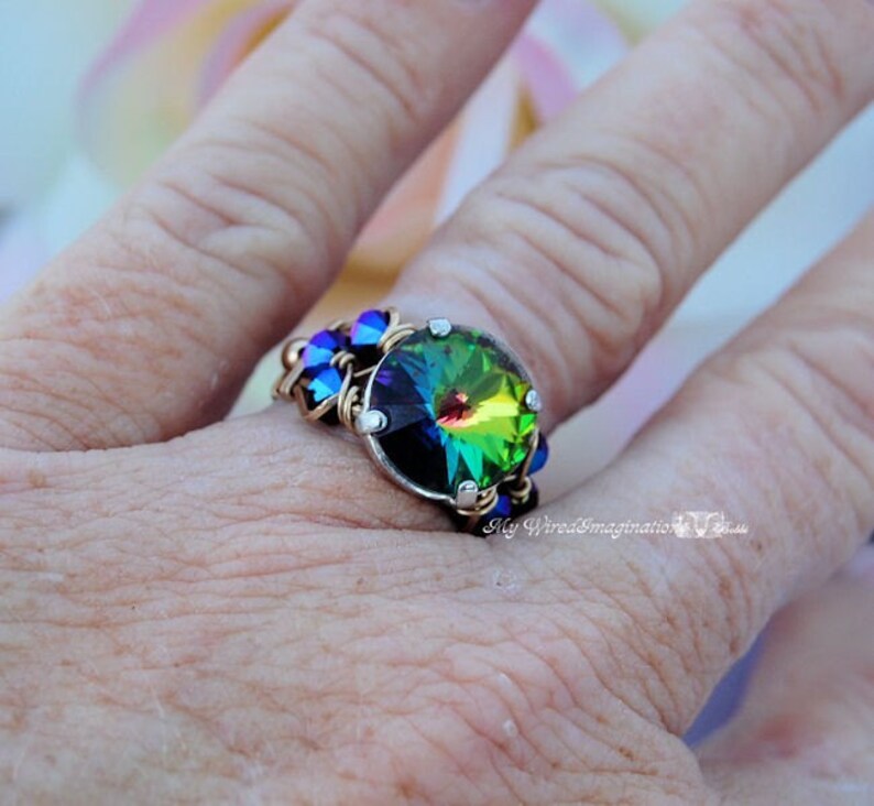 May include: A handcrafted ring featuring a large, iridescent crystal with a rainbow effect. The crystal is set in a silver-toned metal with gold wire accents and smaller blue crystals. The ring is on a finger.