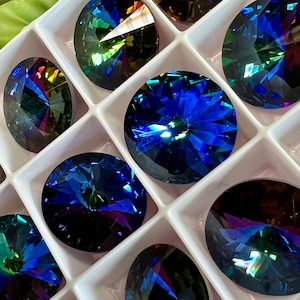 Puede incluir: Primer plano de una variedad de piedras de cristal azul y verde iridiscentes en una vitrina blanca. Las piedras de cristal están dispuestas en una cuadrícula y tienen un diseño facetado.
