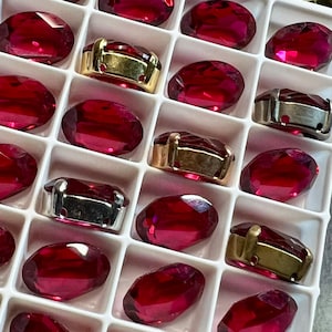 May include: Close-up of a white tray filled with oval-shaped, deep red gemstones. Some gemstones are set in gold-colored metal settings, while others are in silver-colored metal settings. The gemstones are faceted and reflect light.