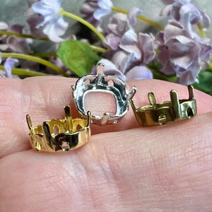 May include: Three metal jewelry settings with prongs for securing gemstones. One setting is silver-toned, one is gold-toned, and one is bronze-toned. Each setting features a circular base with vertical prongs extending upward to hold a stone.