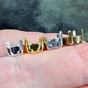 K&ouml;nnte beinhalten: Vier kleine Schmuckfassungen aus Metall in einer Reihe, jede mit vier vertikalen Krappen versehen. Die Fassungen sind in Silber-, Gold-, hellen Silber- und Bronzet&ouml;nen gehalten und dazu bestimmt, einen kleinen Stein oder Cabochon sicher zu halten.
