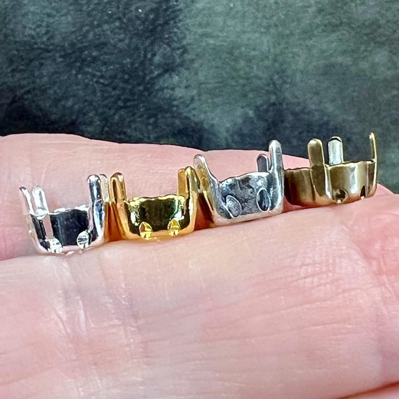 K&ouml;nnte beinhalten: Vier kleine Schmuckfassungen aus Metall in einer Reihe, jede mit vier vertikalen Krappen versehen. Die Fassungen sind in Silber-, Gold-, hellen Silber- und Bronzet&ouml;nen gehalten und dazu bestimmt, einen kleinen Stein oder Cabochon sicher zu halten.