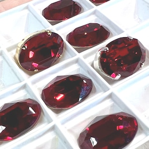 Puede incluir: Primer plano de varias piedras preciosas rojas oscuras facetadas, de forma ovalada. Algunas están engastadas en biseles de oro o plata, mientras que otras están sueltas. Las gemas están dispuestas en una bandeja blanca.