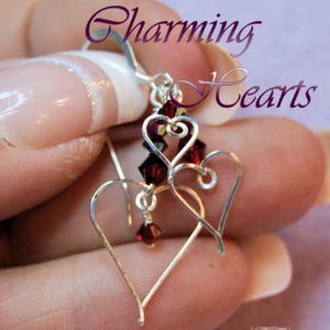 Könnte beinhalten: Ein Paar Ohrringe aus silberfarbenem Draht mit zwei herzförmigen Umrissen und dunkelroten facettierten Perlen. Die Ohrringe hängen an silberfarbenen Haken und zeigen den Text Charming Hearts in einer violetten Schreibschrift.