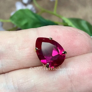 May include: A single, pear-shaped, bright pink rhinestone with gold-toned settings. The rhinestone is on a finger.