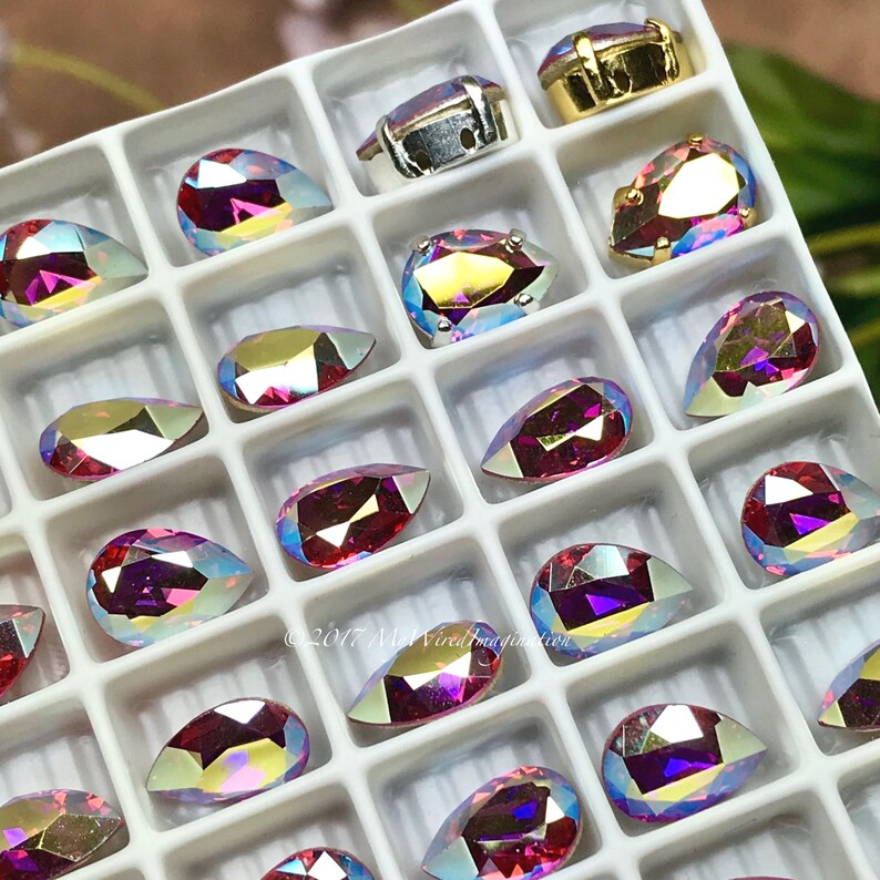 Puede incluir: Una bandeja de pedrer&iacute;a iridiscente en varias formas, incluyendo l&aacute;grimas y &oacute;valos. La pedrer&iacute;a tiene un efecto brillante multicolor con tonos de rosa, morado y dorado. Algunos est&aacute;n engastados en engastes de metal dorado o plateado.