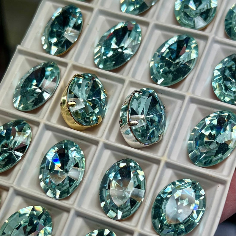 May include: A close-up of a tray filled with oval-shaped, light blue rhinestones. The rhinestones are arranged in rows and are set in a white tray.
