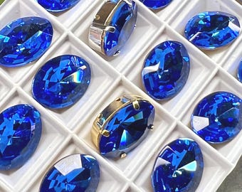 May include: An assortment of vibrant blue oval-cut gemstones and two gold-plated settings are arranged in a white grid. The gemstones have multiple facets, reflecting light. The gold settings are rectangular with rounded edges.