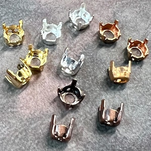 May include: A collection of small metal prong settings for gemstones, featuring various metallic finishes including gold, silver, copper, and bronze tones. Each setting has a circular base with four upright prongs designed to hold a stone securely.