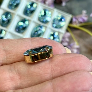 May include: A single, clear, faceted, light blue gemstone with a gold setting. The gemstone is in the shape of a rectangle with rounded corners. The gemstone is set in a gold frame with two holes on the bottom.