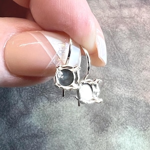 May include: A pair of silver-toned earrings with square, bezel-set gemstones. The gemstones have a light blue-gray hue. The earrings are held up by a person's hand, showcasing the intricate design and craftsmanship.