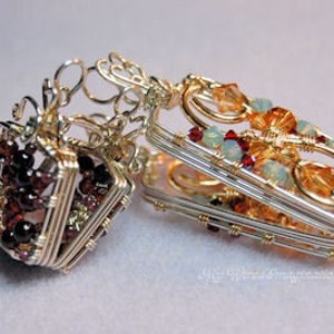 以下が含まれることがあります： 赤と透明な宝石が付いた、銀と金のワイヤーで包まれた2つのペンダント。左側のペンダントは立方体で、右側のペンダントは長方形です。