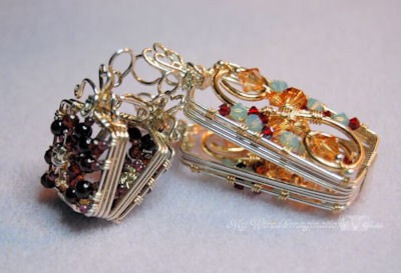 以下が含まれることがあります： 赤と透明な宝石が付いた、銀と金のワイヤーで包まれた2つのペンダント。左側のペンダントは立方体で、右側のペンダントは長方形です。