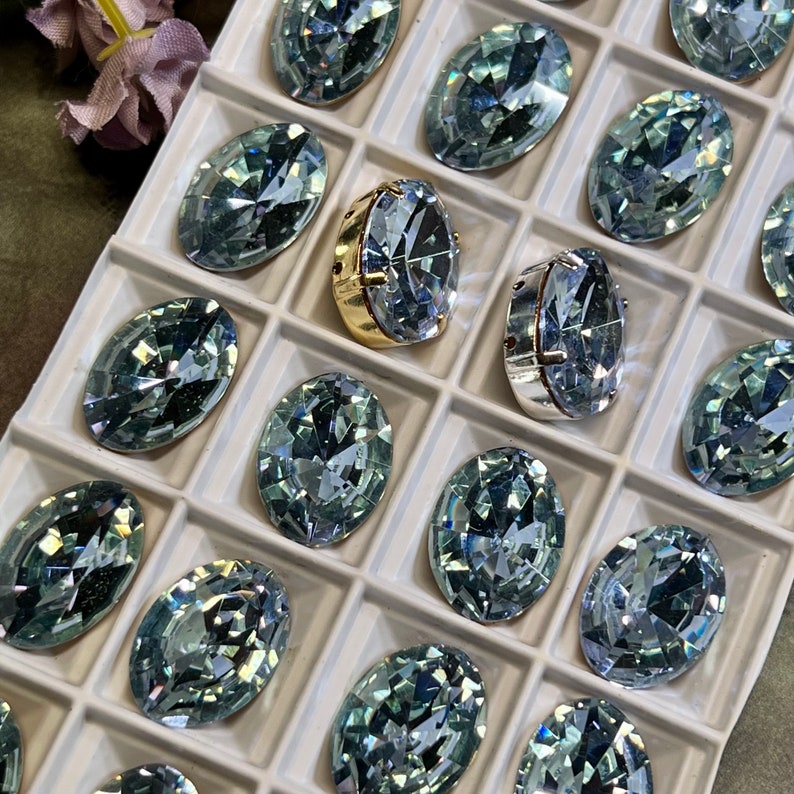 May include: A tray of oval-shaped, light blue rhinestones with a gold and silver setting. The rhinestones are arranged in a grid pattern.