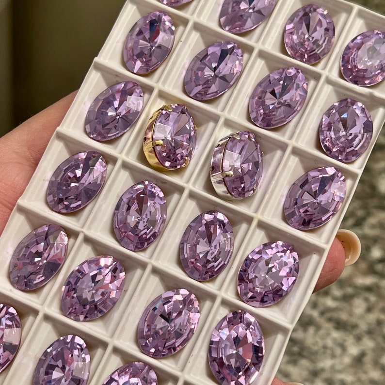 May include: A close-up of a white tray filled with small, oval-shaped, light purple rhinestones. The rhinestones are arranged in rows and columns.