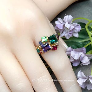 May include: A gold-toned ring featuring a cluster of multi-colored square-cut gemstones including purple, green, and red stones. The band is constructed from twisted wire metal. The text "©2018 MyWiredImagination" is visible at the bottom.